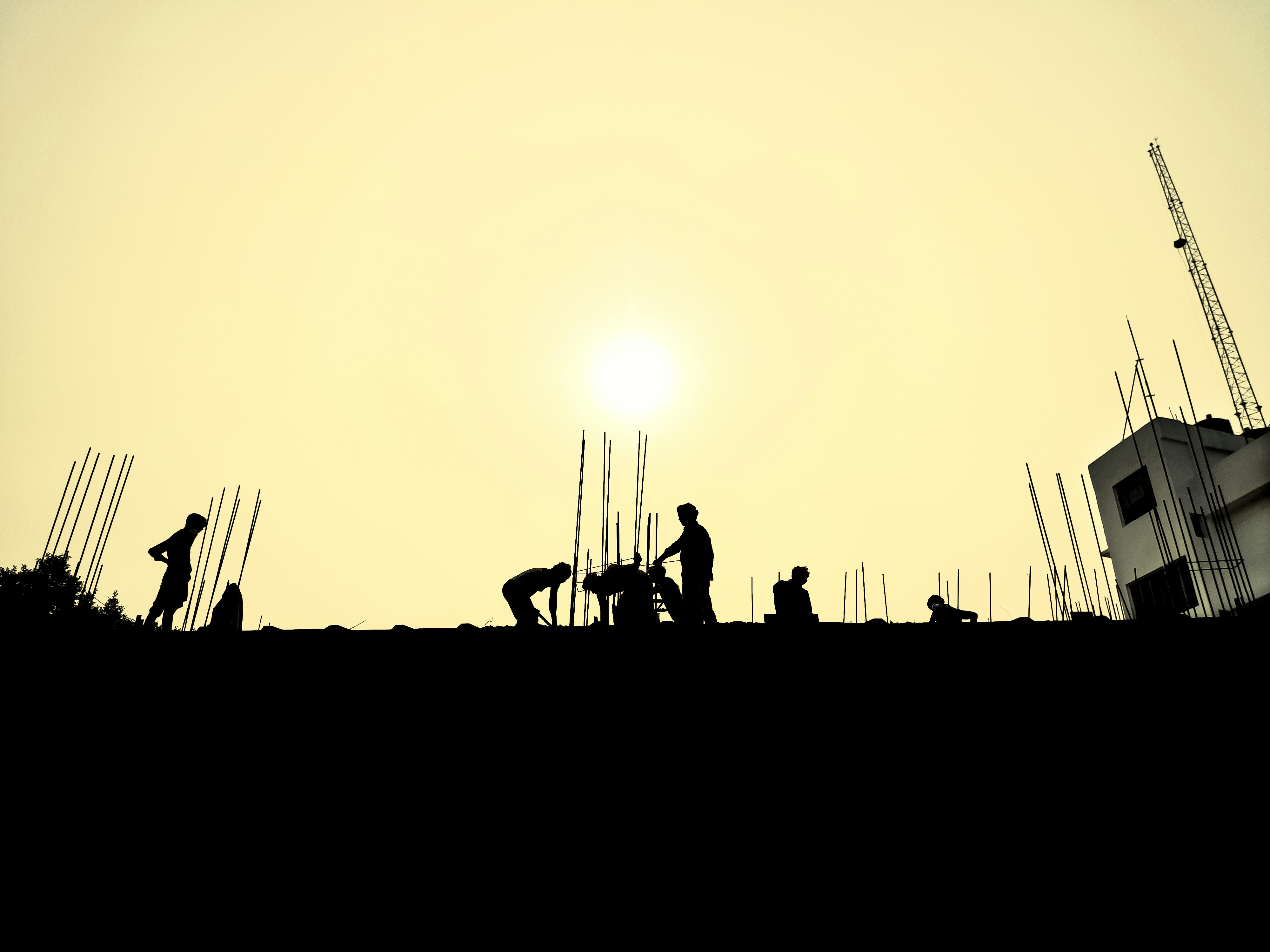 Silhouettes of a few constructions workers laboring on the foundational parts of the building in the sun making the sky light yellow